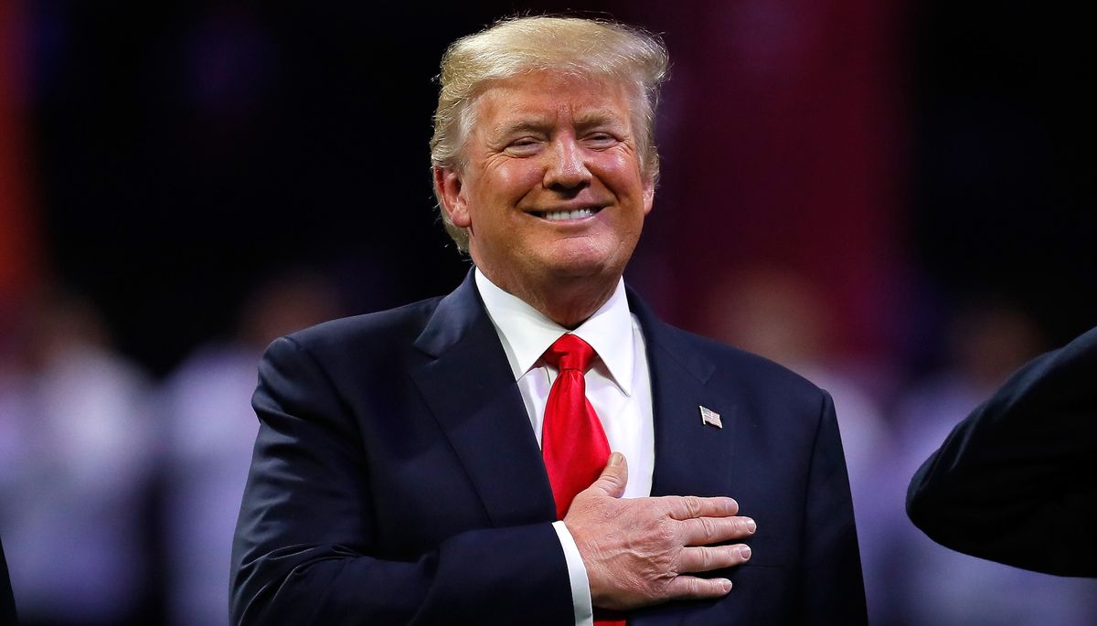 Donald Trump in a dark suit with a red tie, hand placed over his heart.