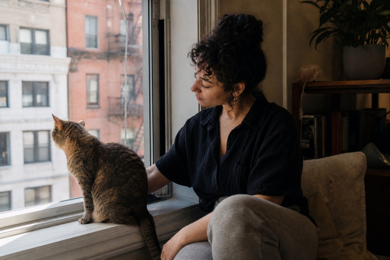 Person sitting by a window beside a cat on the sill in a softly lit apartment
