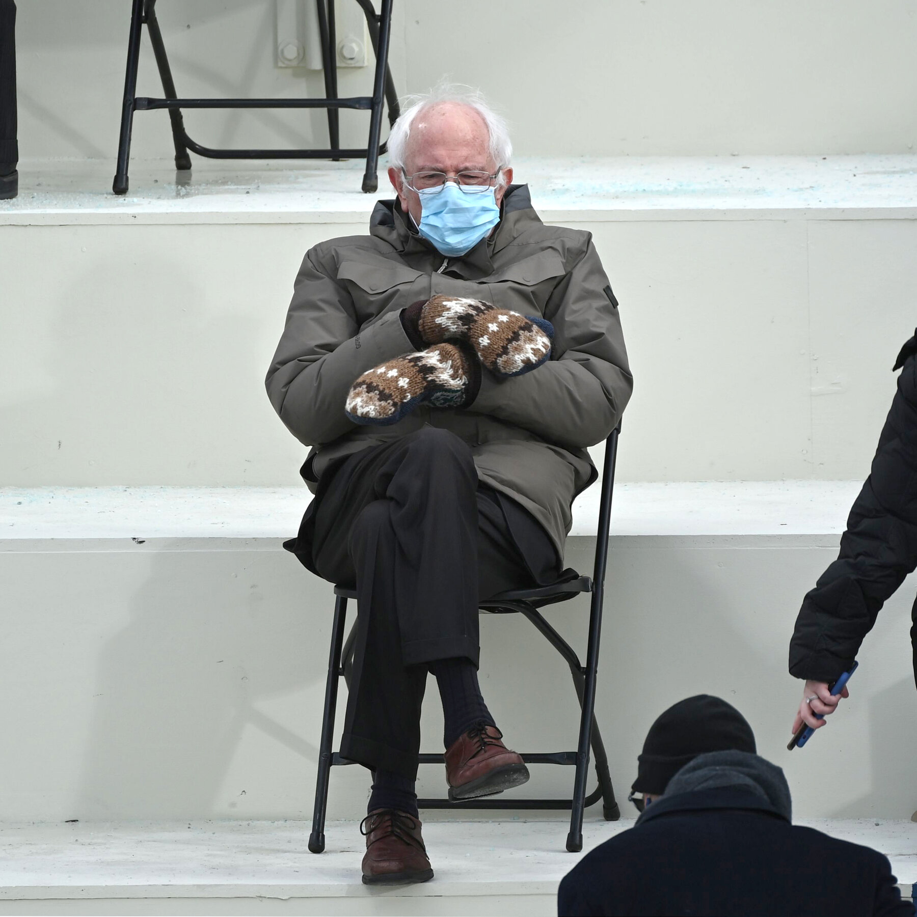 Bernie Sanders sitting on a folding chair at an inauguration, wearing a mask and mittens.