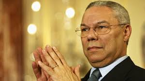 Portrait of Colin Powell with clasped hands and glasses against a warm blurred background.