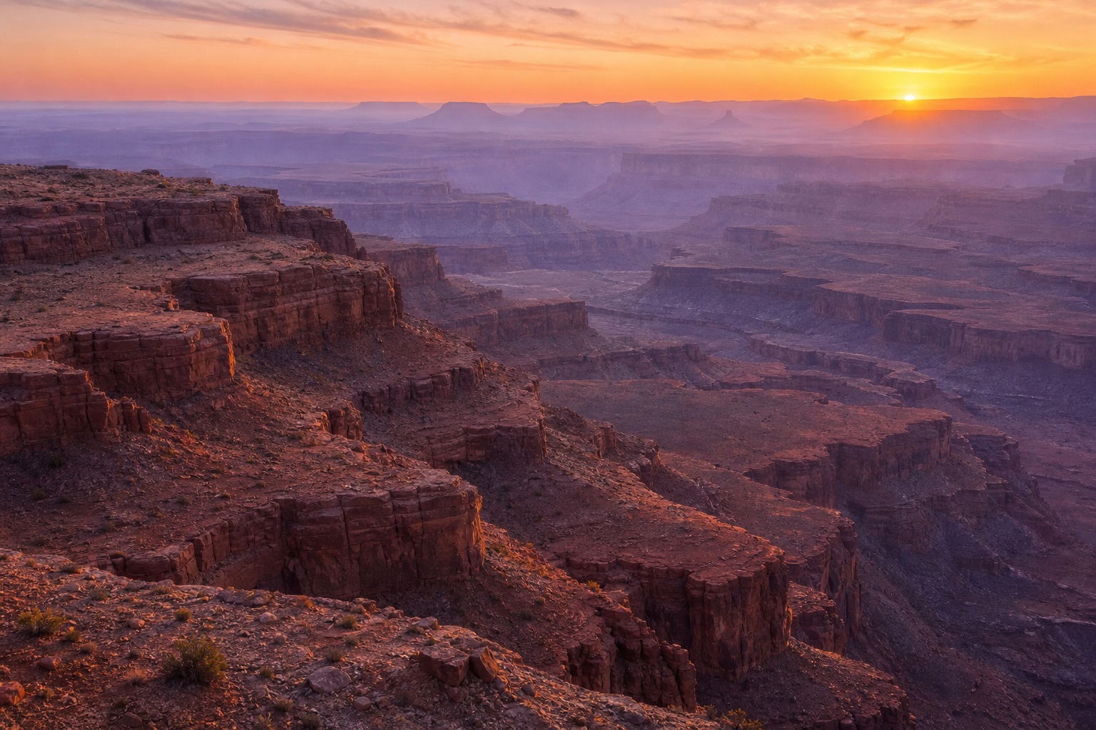 Sunrise over layered sandstone canyon terraces in a vast desert landscape.
