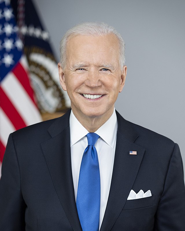 Smiling portrait of President Joe Biden in a dark suit and blue tie before a U.S. flag