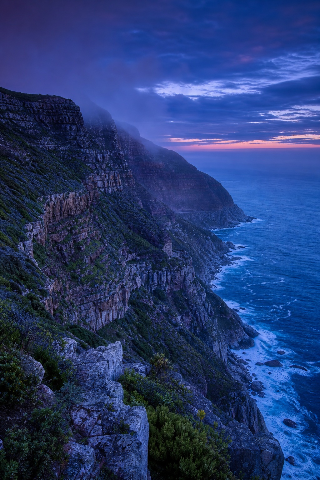 Steep rocky coastal cliffs beside the ocean under a blue hour sky.