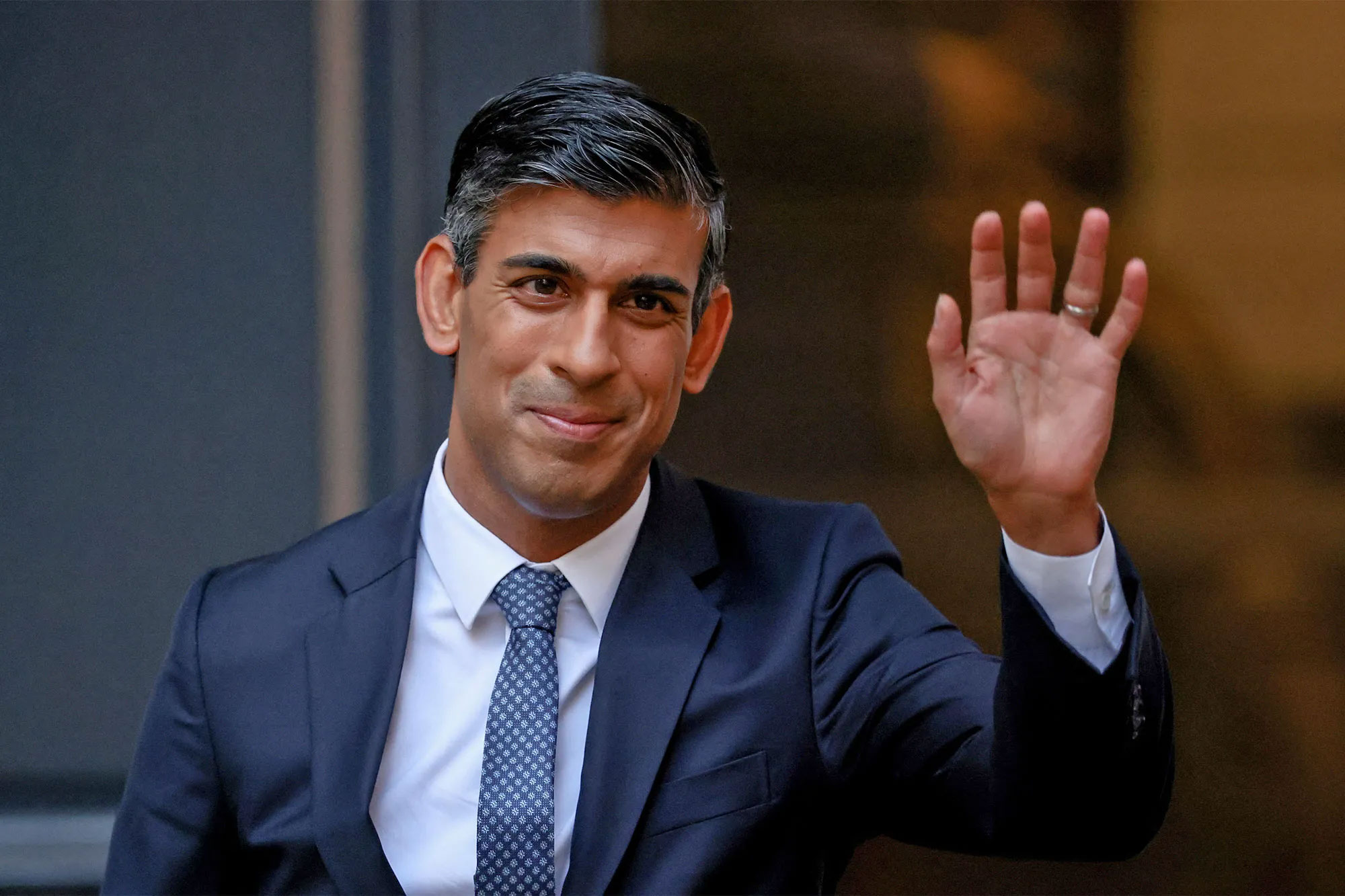 Rishi Sunak in a suit waving with one hand against a dark background