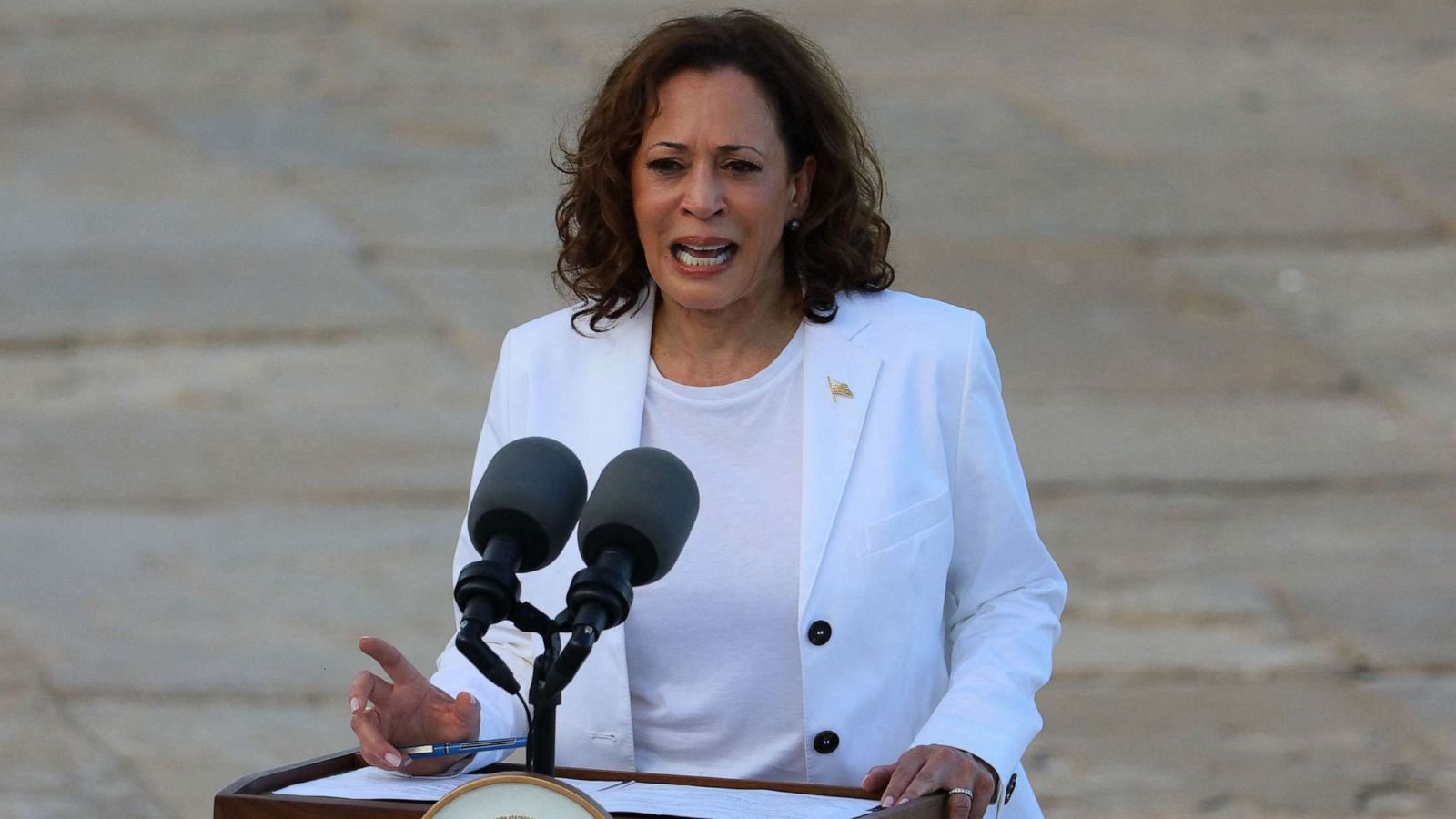 Woman in a white blazer speaking at a podium with microphones.