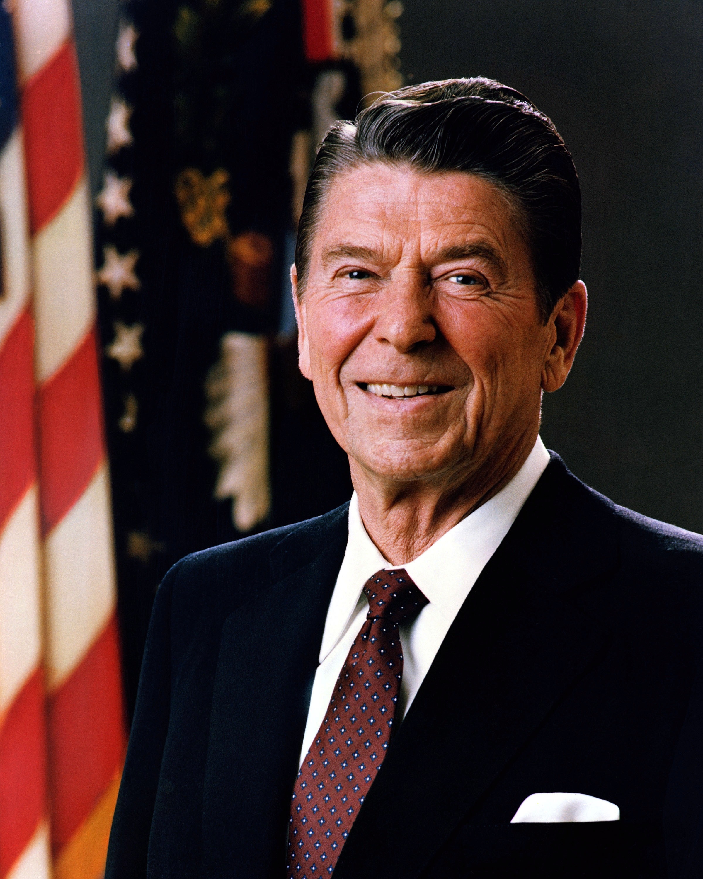 Formal portrait of Ronald Reagan smiling in a suit with an American flag behind him.