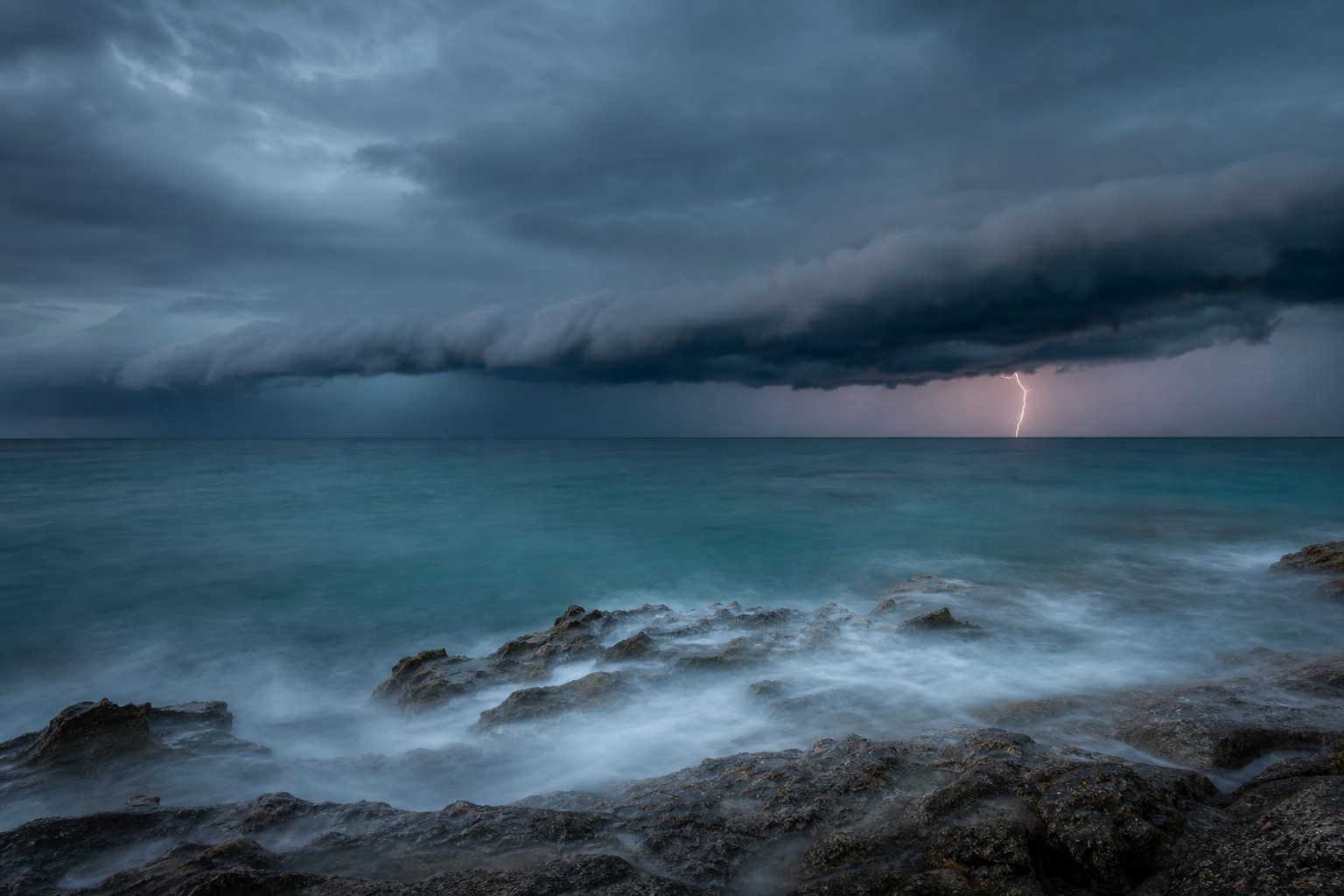 Dark storm clouds over a rough ocean with lightning near the horizon and rocky shoreline.