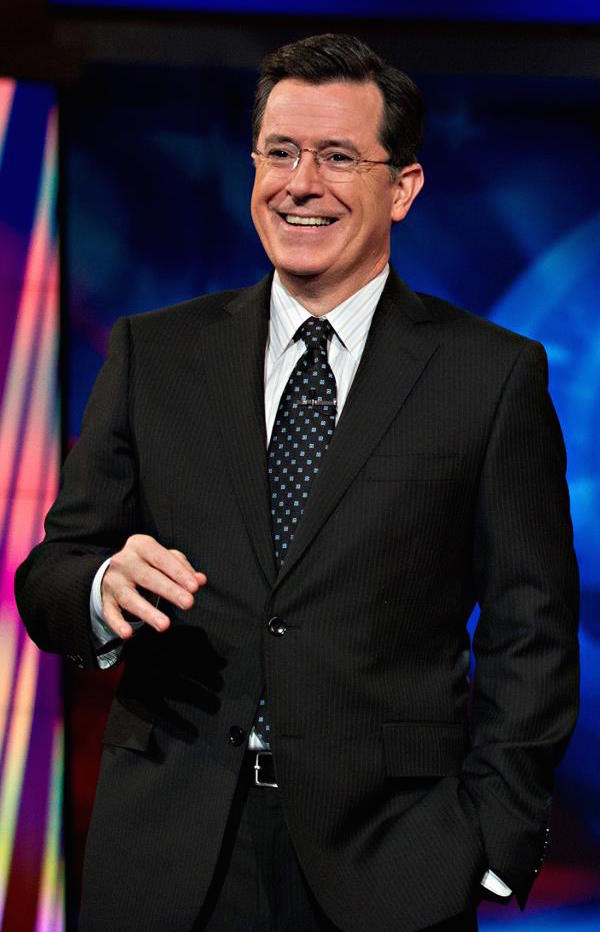 Stephen Colbert smiling in a suit on a brightly lit stage