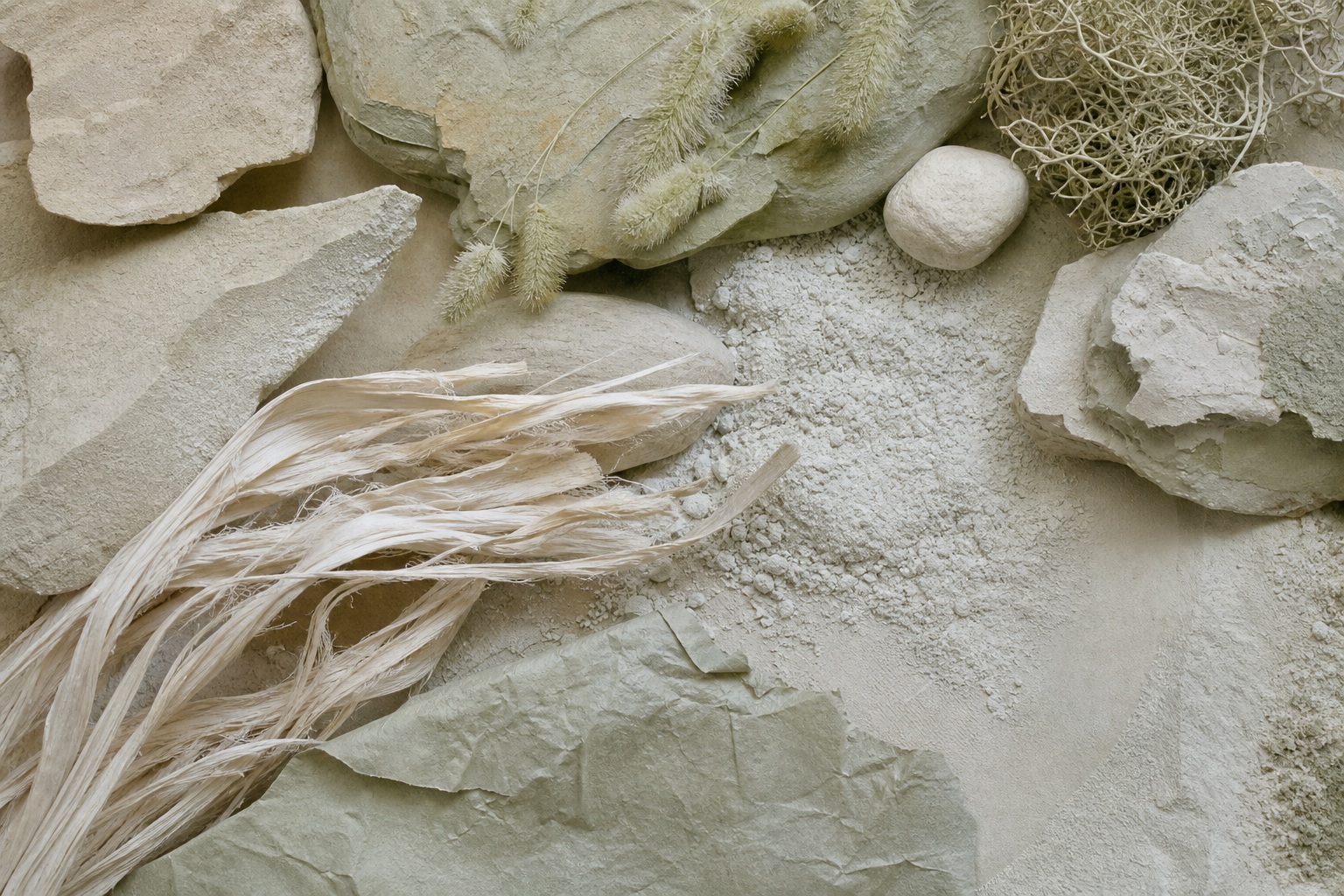 Neutral ceramic and stone fragments arranged on a textured sage-toned surface with dried stems.