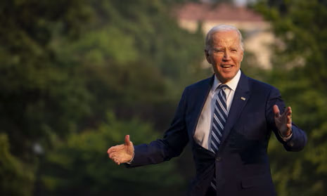 Joe Biden standing outdoors in a suit with blurred greenery behind him.