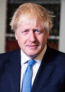 Boris Johnson in a dark suit and blue tie against a blurred indoor background.