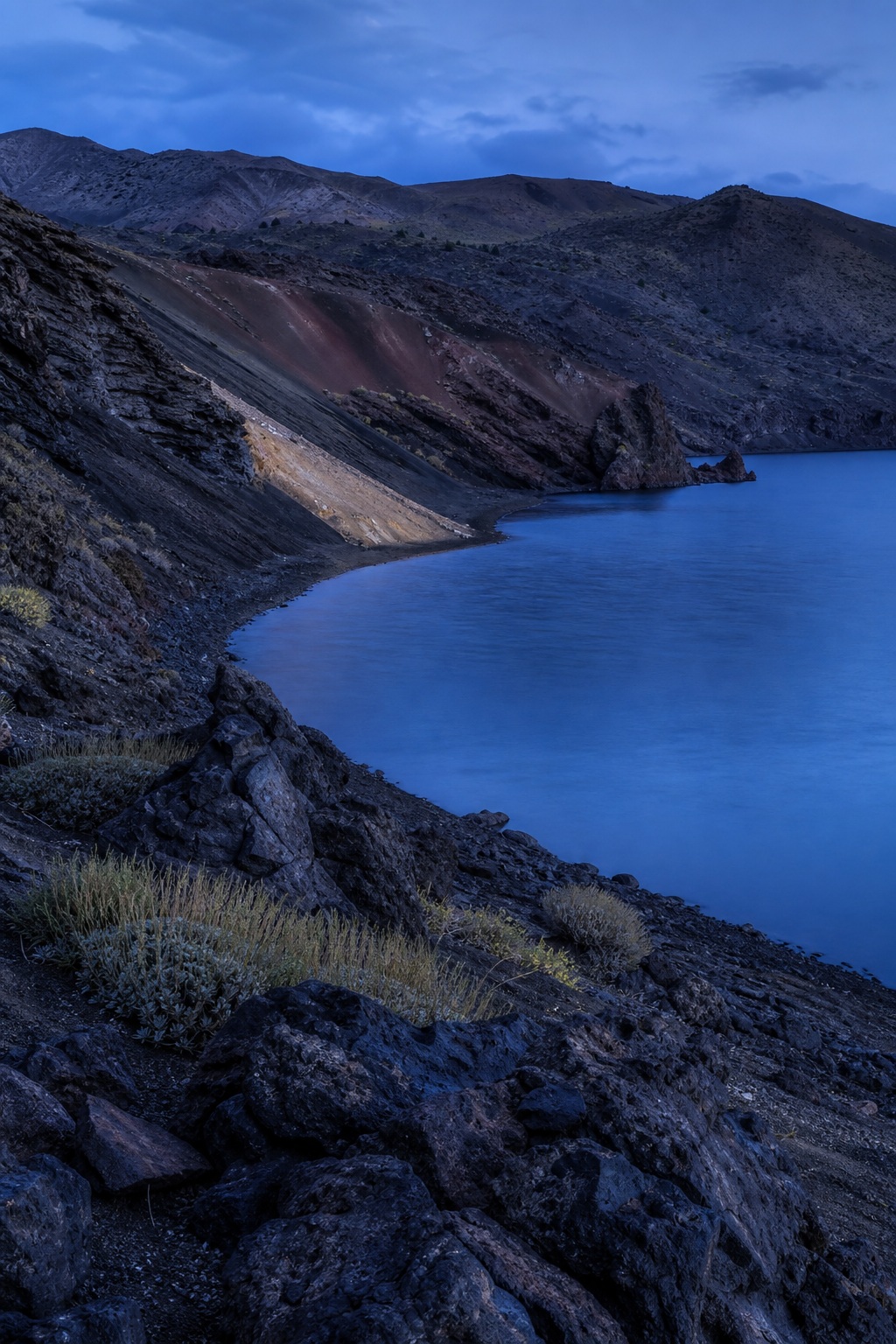 Dark volcanic rocks along a blue caldera lake shore at blue hour