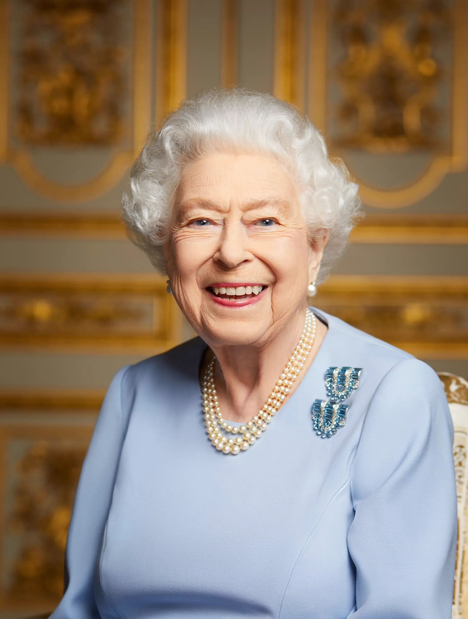 Queen Elizabeth II smiling in a light blue formal dress and pearl necklace.