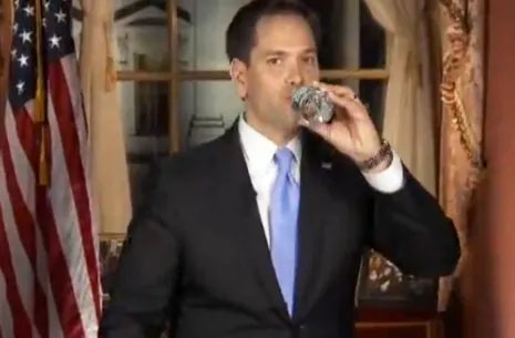 A man in a suit drinking from a mug in an office setting with flags behind him.