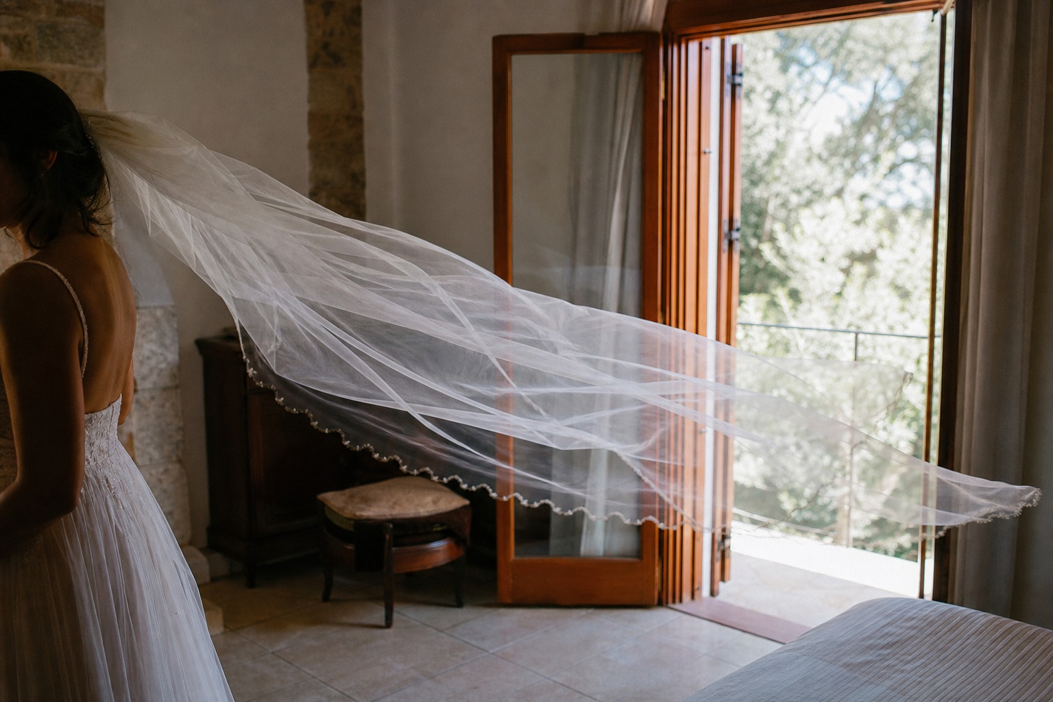 Bride standing indoors with a long veil flowing toward an open doorway.