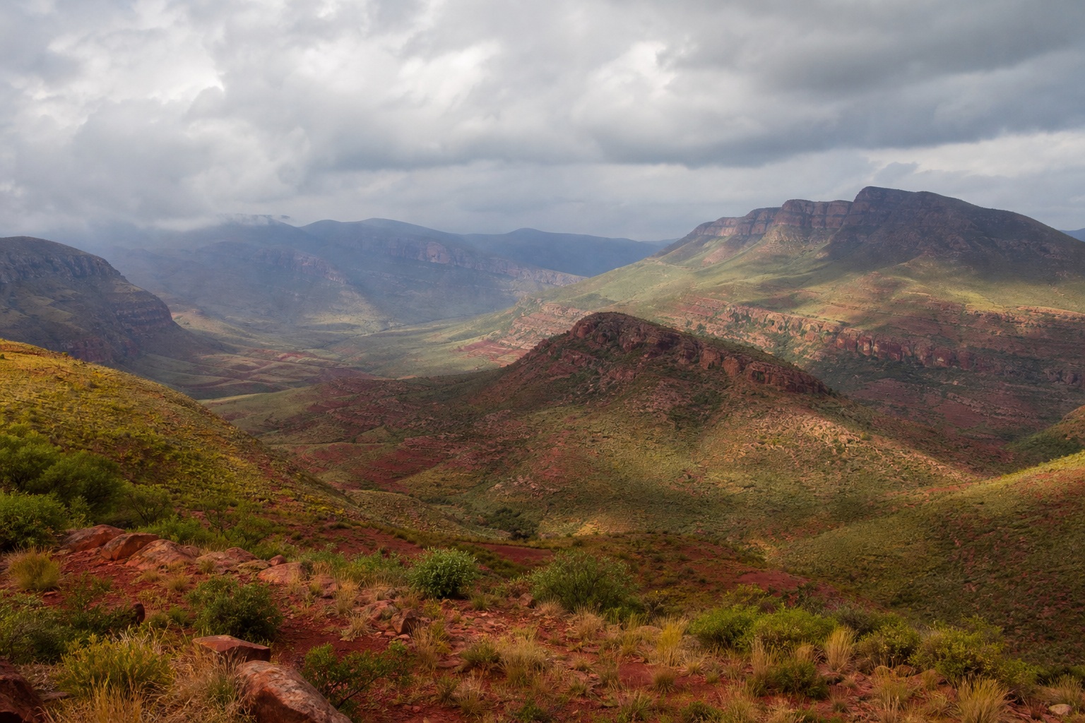 Red earth valley with rolling hills, distant mountains, and cloudy skies.