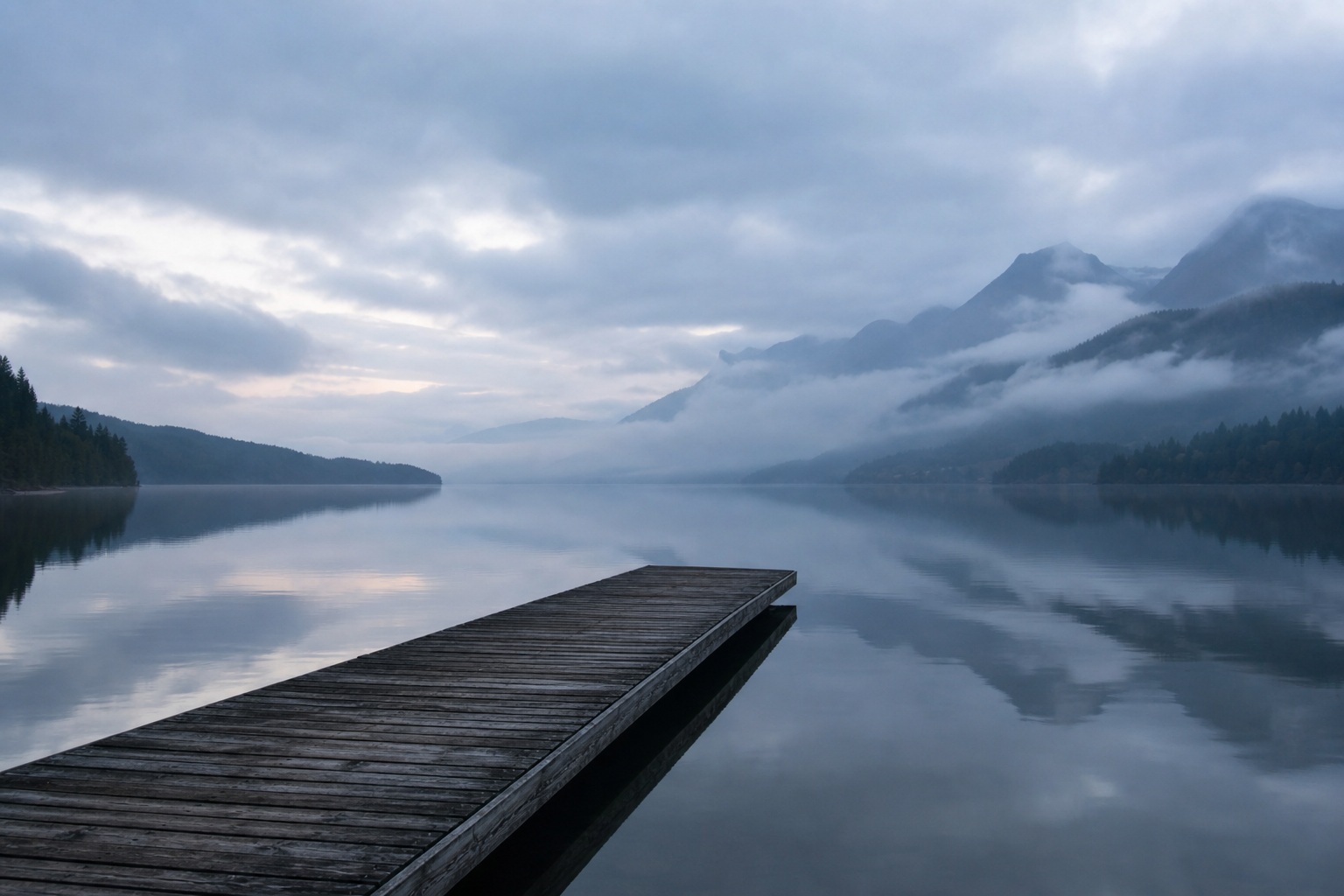 Wooden dock extending into a misty lake with mountains reflected in calm water