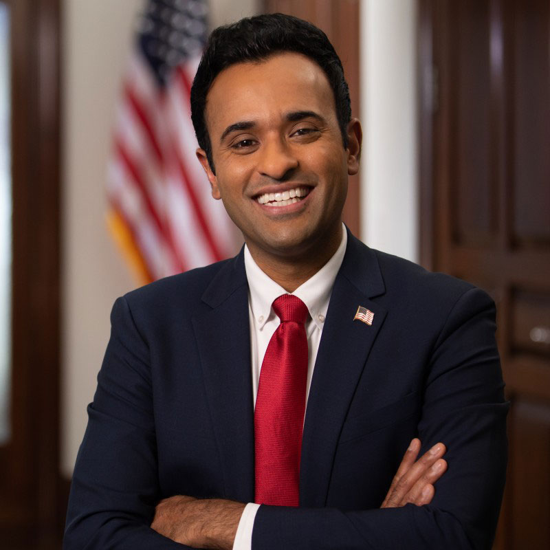 Smiling man in a dark suit and red tie standing with arms crossed in front of a flag.