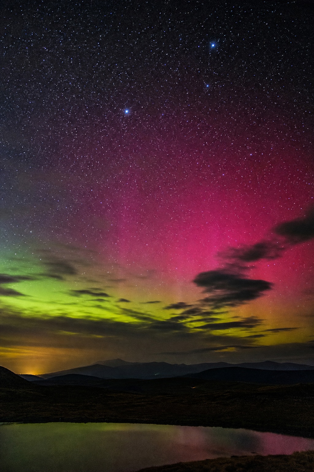 Colorful starry night sky with green and pink airglow above dark mountains and reflection.
