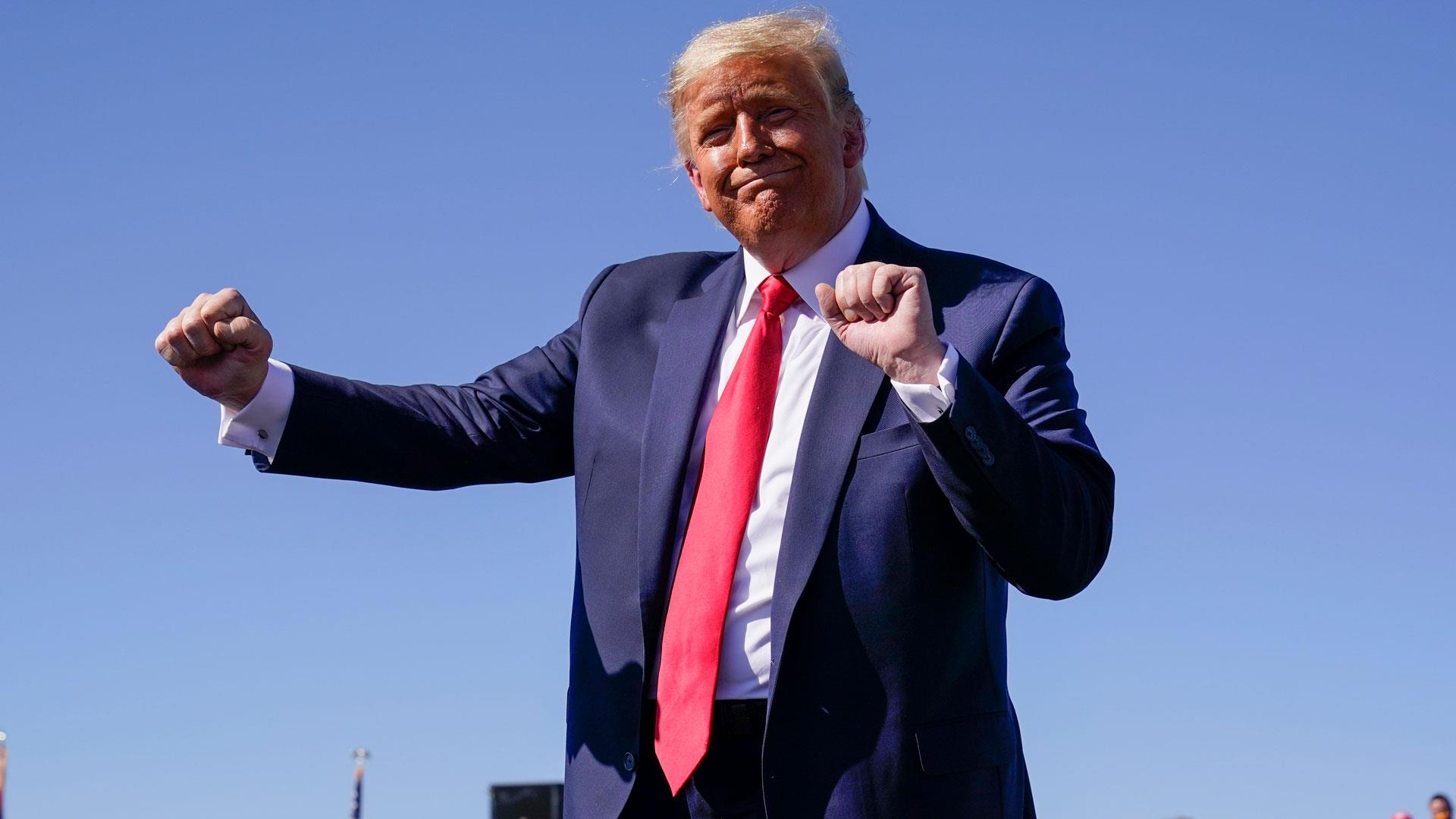 Donald Trump standing outdoors with arms raised, wearing a dark suit and red tie.