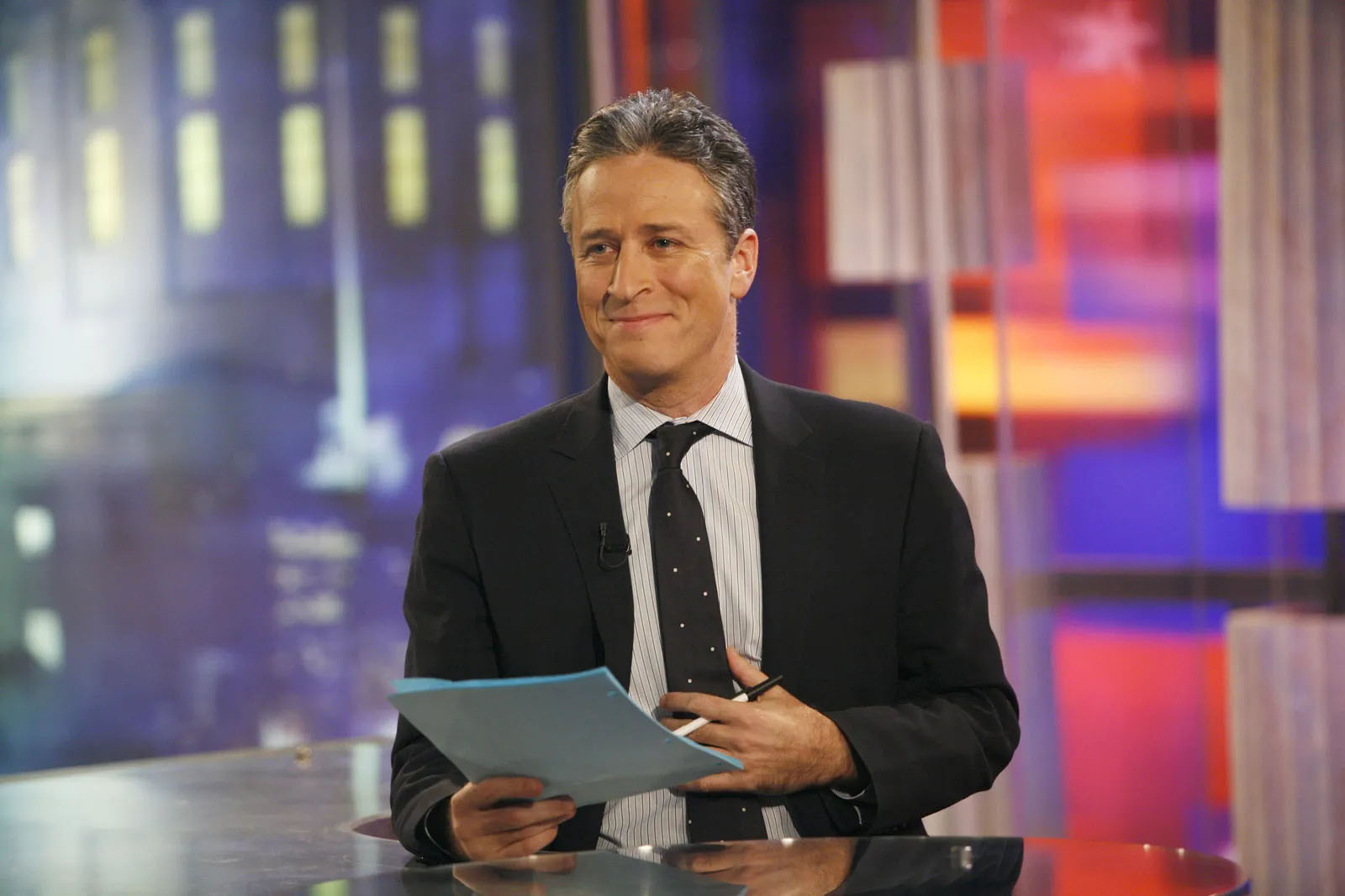 News anchor in a studio reading from a tablet behind a desk