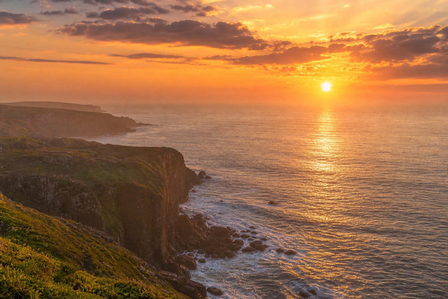 Sunset over the ocean beside a grassy cliff with glowing reflections on the water