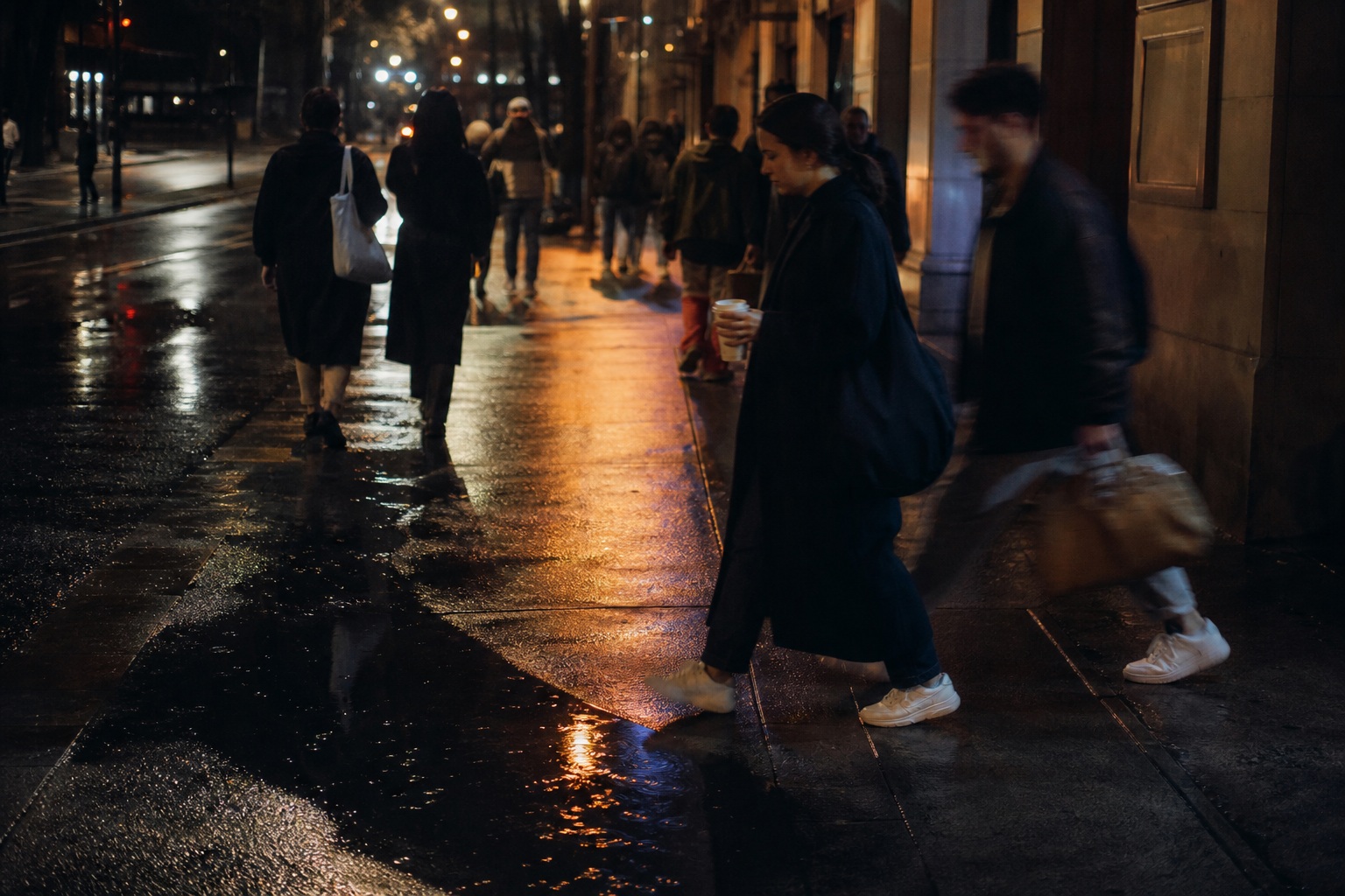 People walking along a rainy city sidewalk at night with wet pavement reflections.