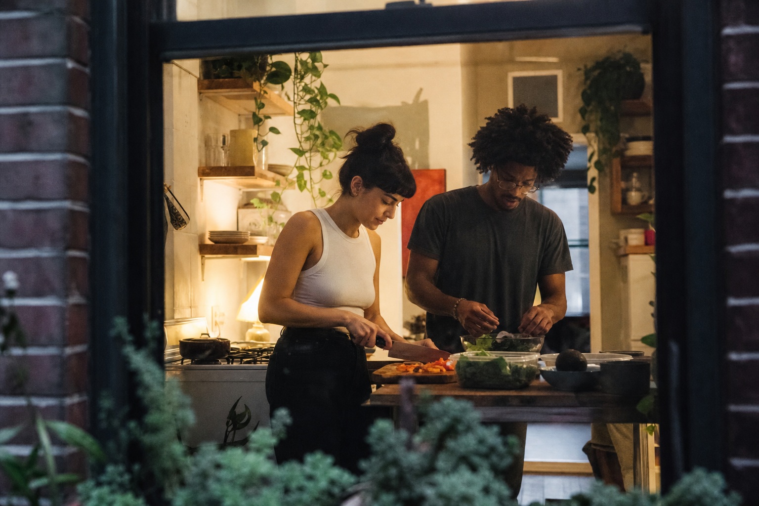 Two people preparing dinner in a kitchen beside a bright apartment window.