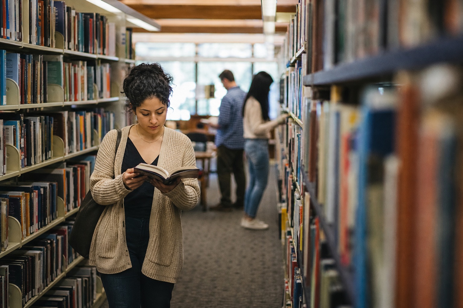Person reading a book in a library aisle with blurred shelves and people behind