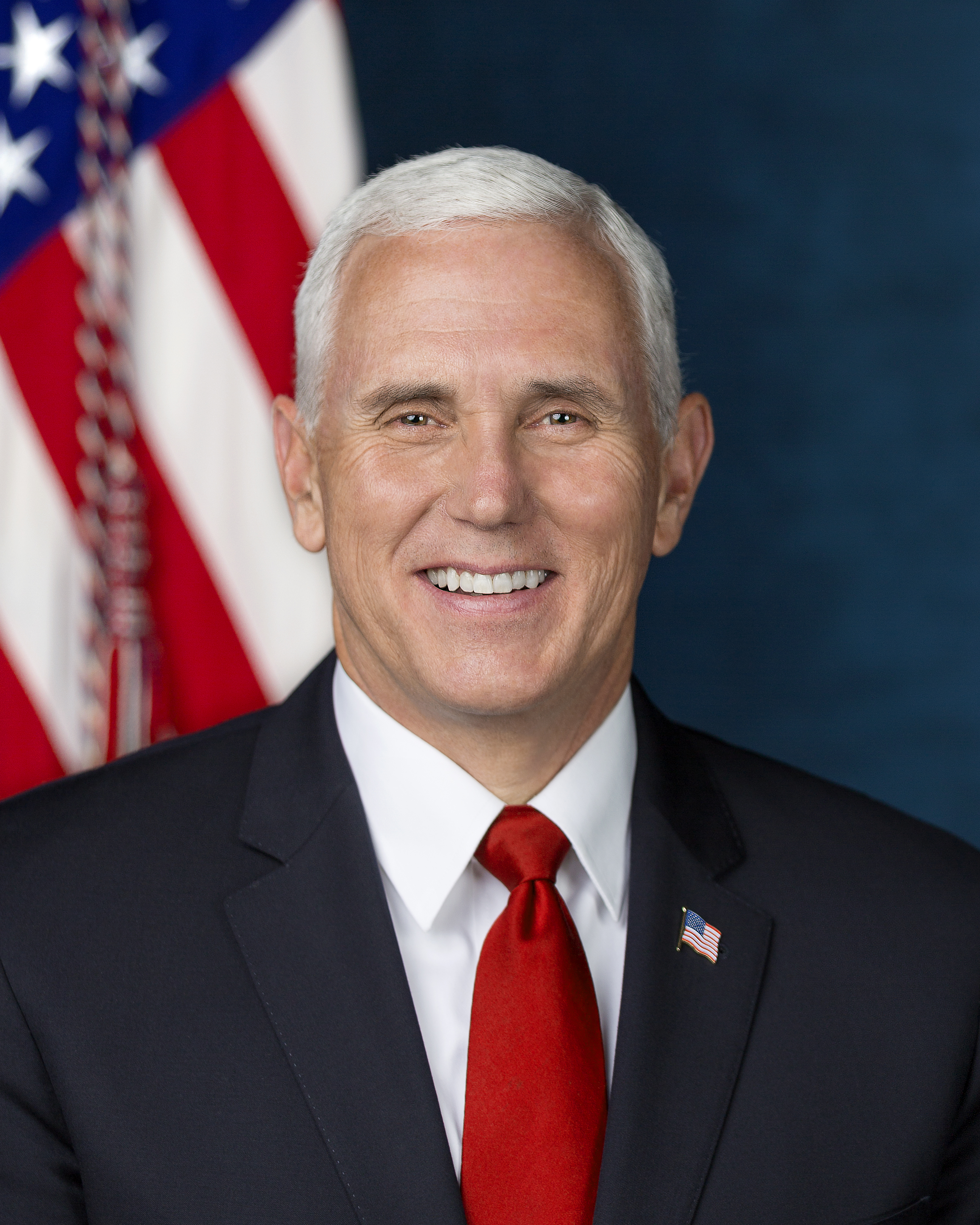 Smiling man in a dark suit, white shirt, and red tie before an American flag.