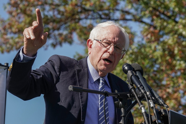 Bernie Sanders speaking at an outdoor podium with microphones, gesturing emphatically with his right hand.