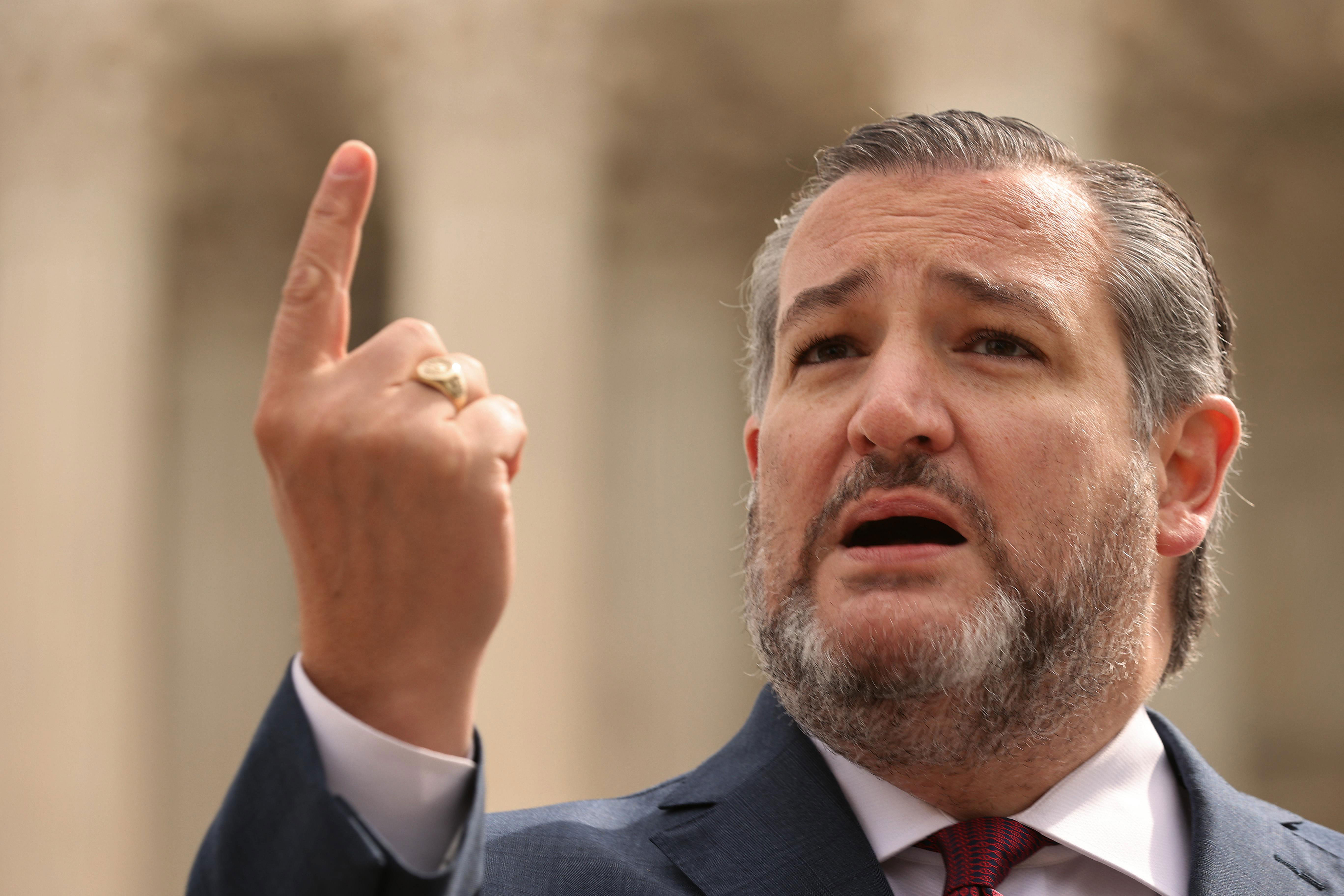 Ted Cruz speaking at an event with one hand raised and a serious expression.