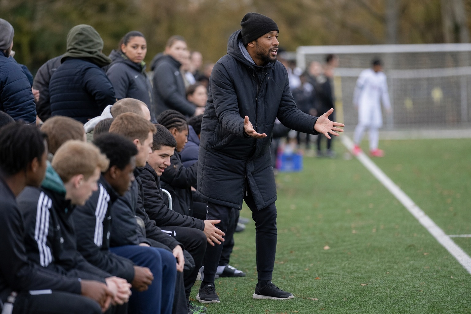 Coach gesturing on a soccer sideline while players sit on the bench beside the field.