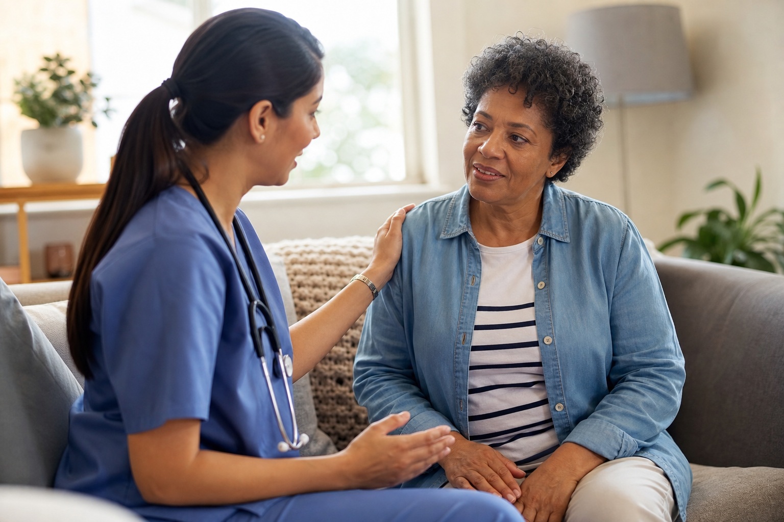 Caregiver in blue scrubs talking with a seated woman in a living room