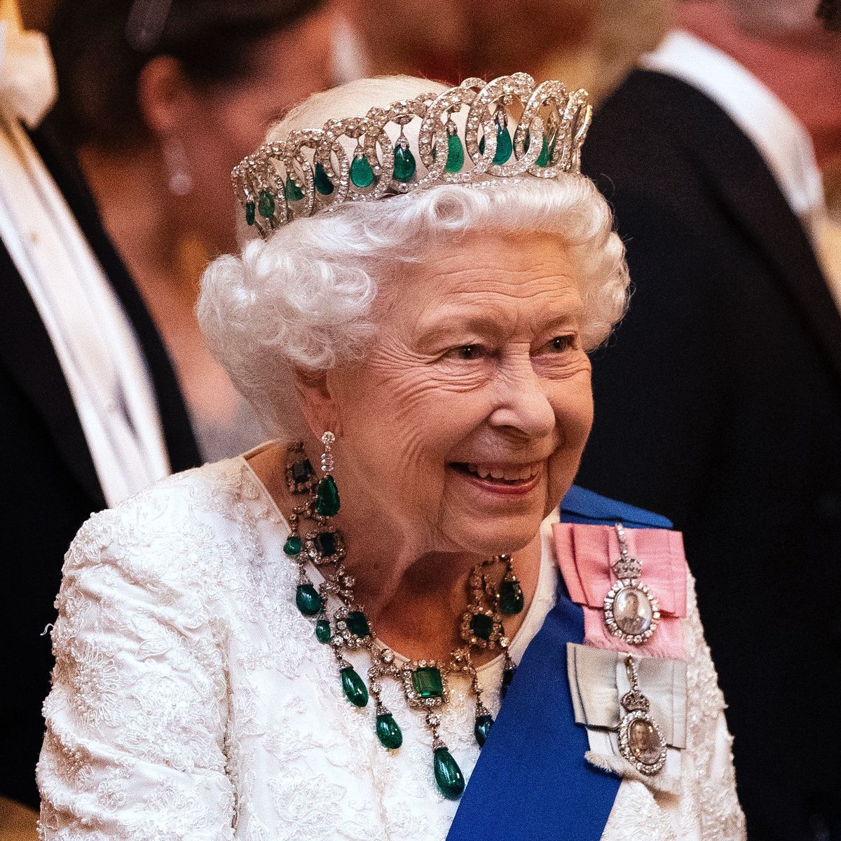 Queen Elizabeth II wearing a crown, white dress, and green jeweled necklace