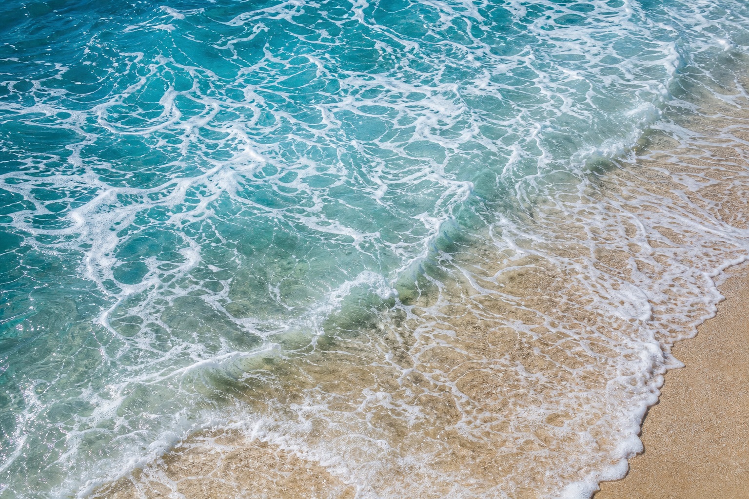 Aerial view of blue ocean waves and white foam over sandy shoreline.