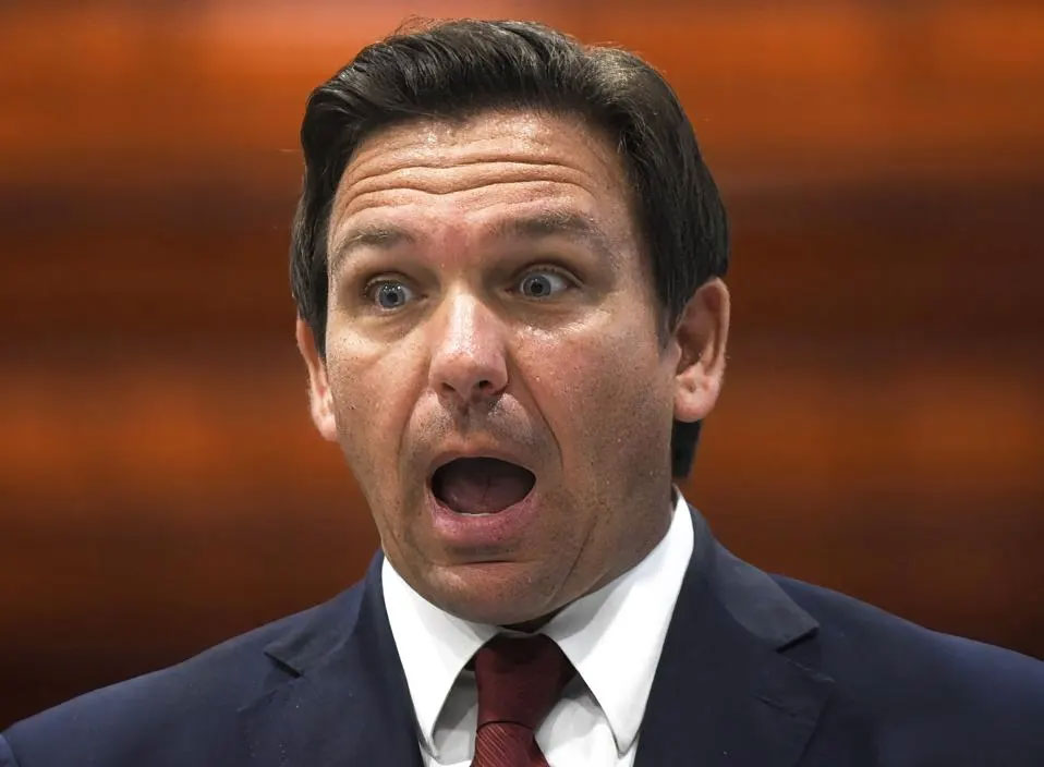 Man in a suit speaking with an open mouth against a blurred warm background.