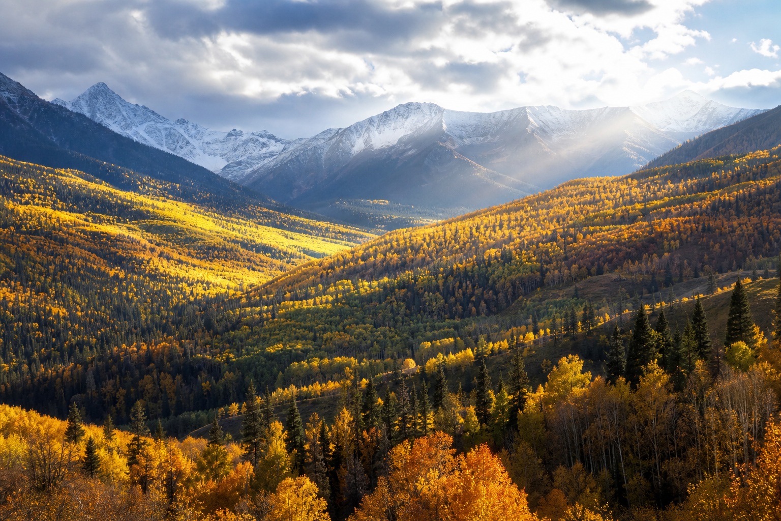 Sunlit autumn valley with golden forest, snowy mountains, and rolling hills