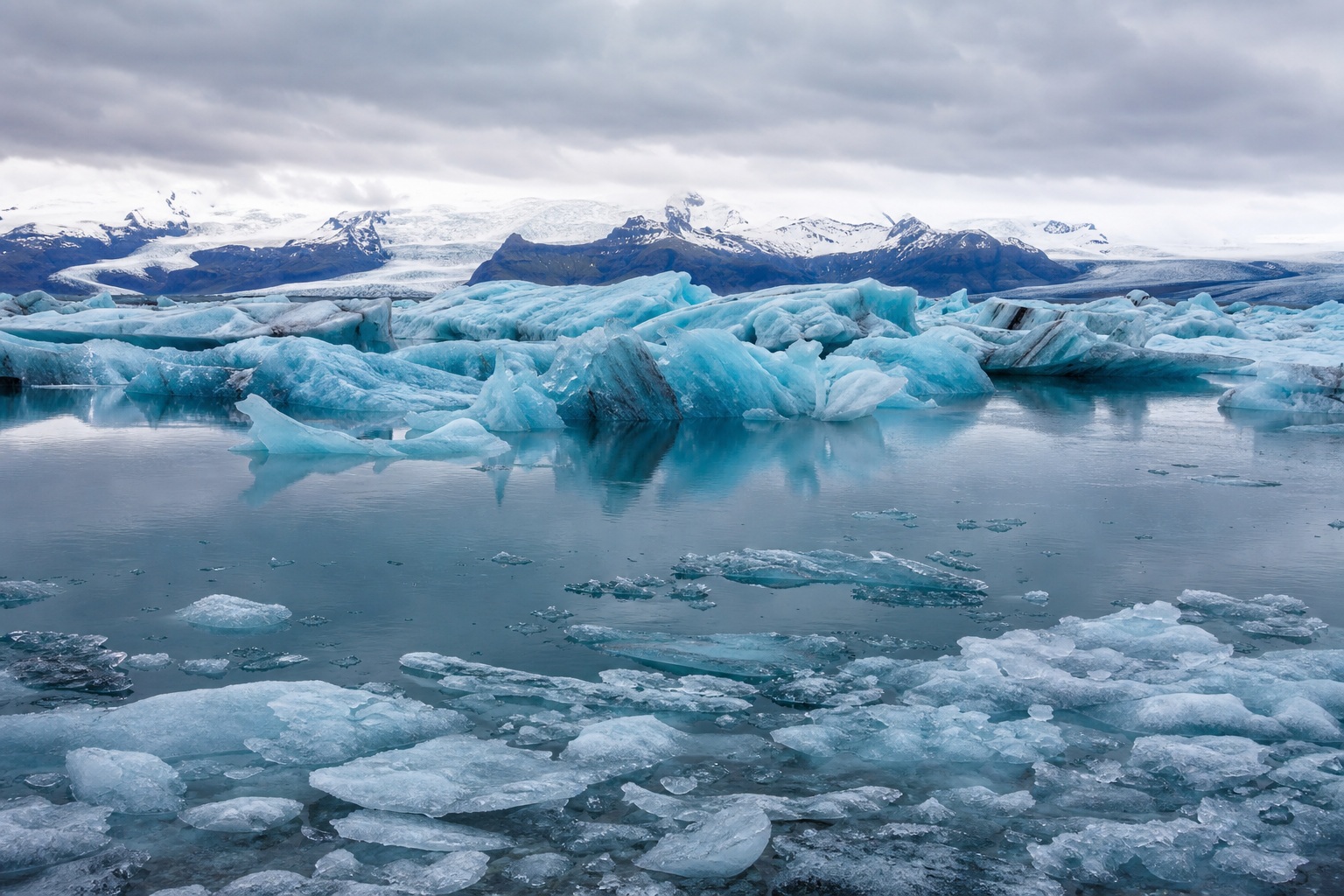 Blue icebergs floating in a frozen lagoon with snowy mountains under a cloudy sky