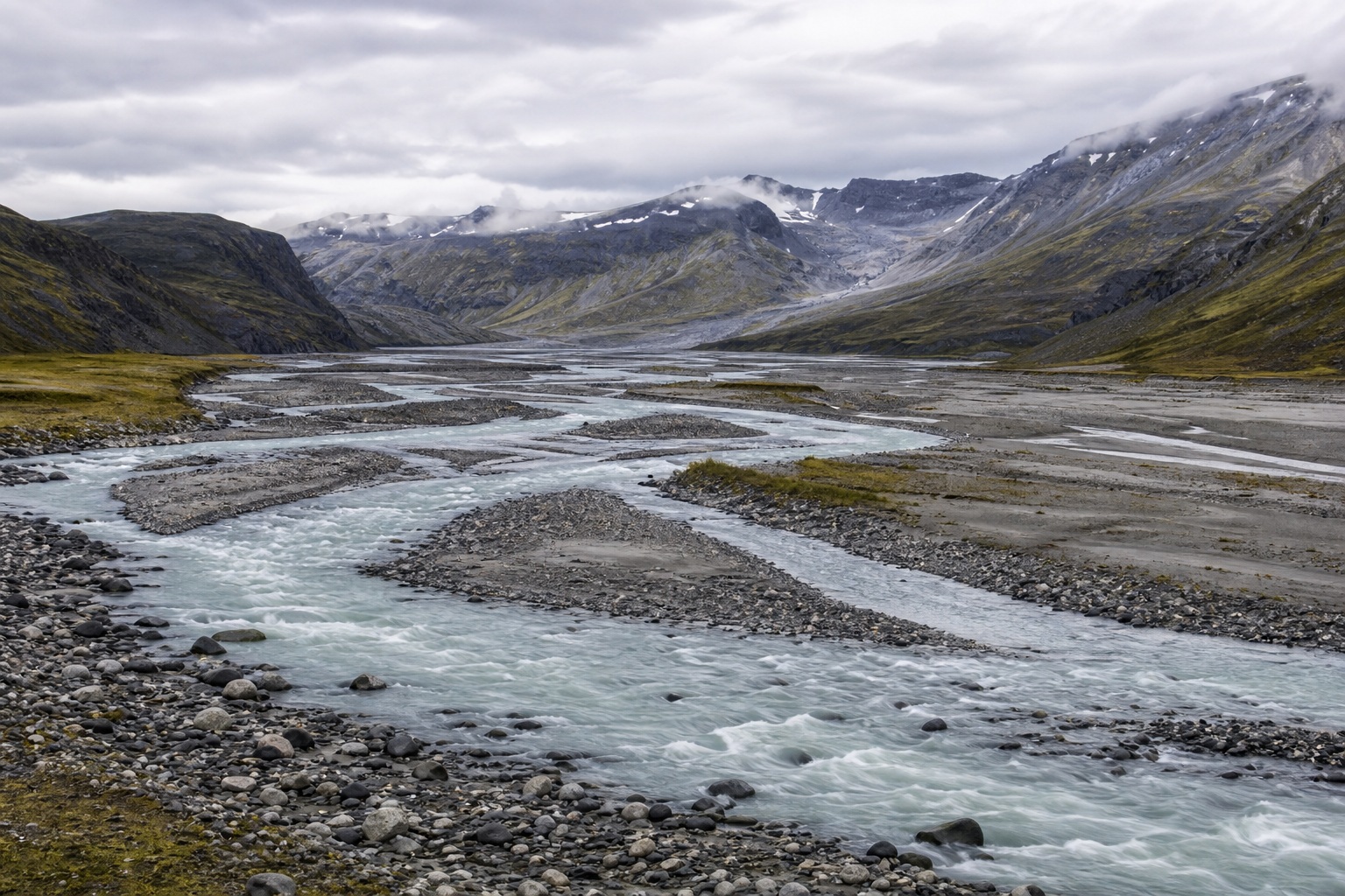 Braided glacial river channels winding through a mountain valley under bright overcast skies.