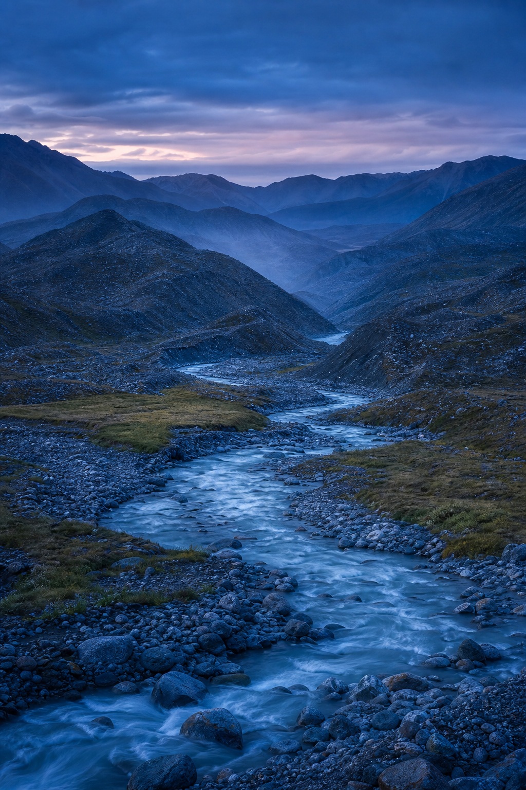 A blue hour glacial valley with a winding river between dark layered mountains.