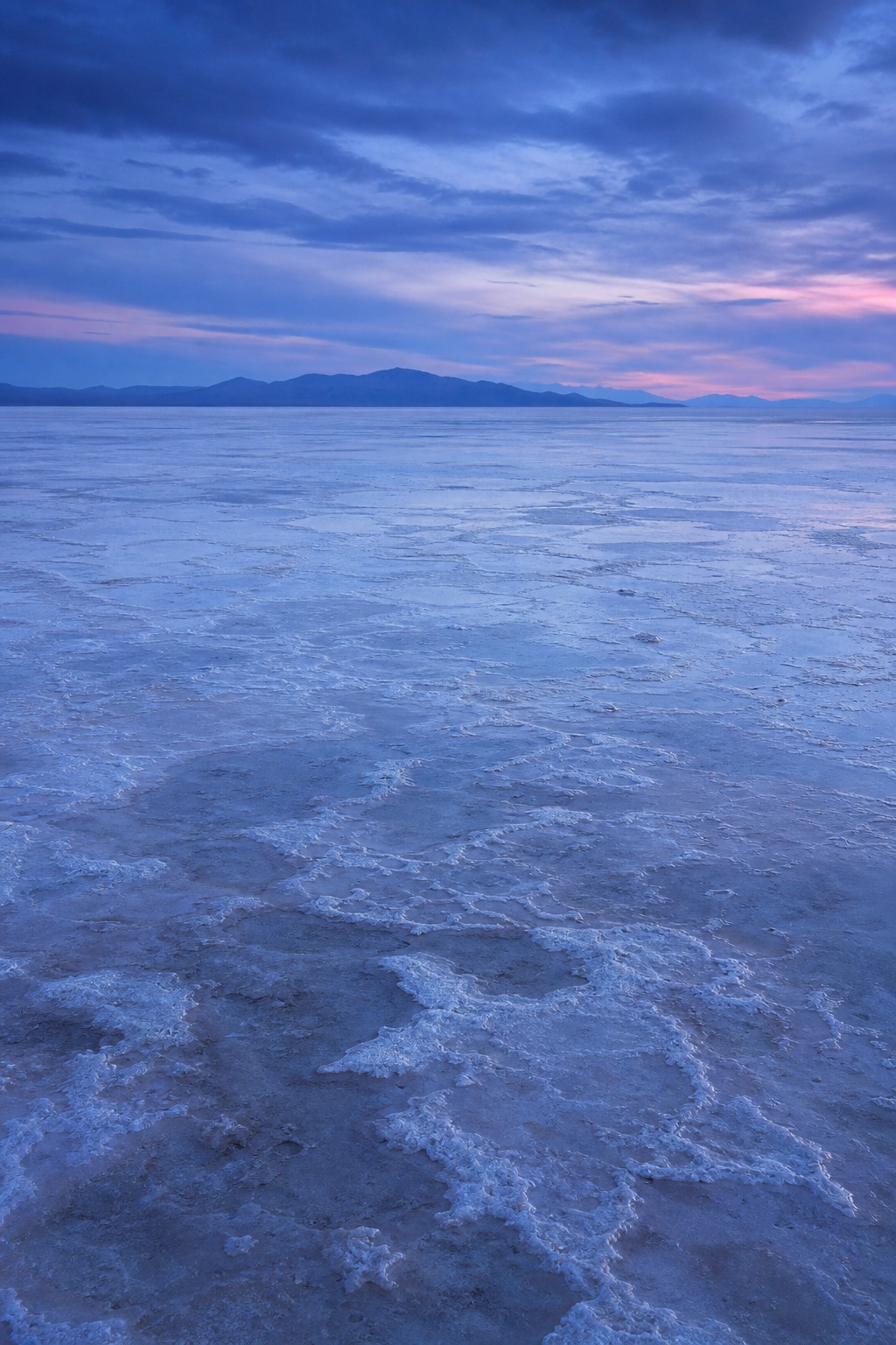 Blue twilight over a salt flat with pale mineral patterns and distant mountains.
