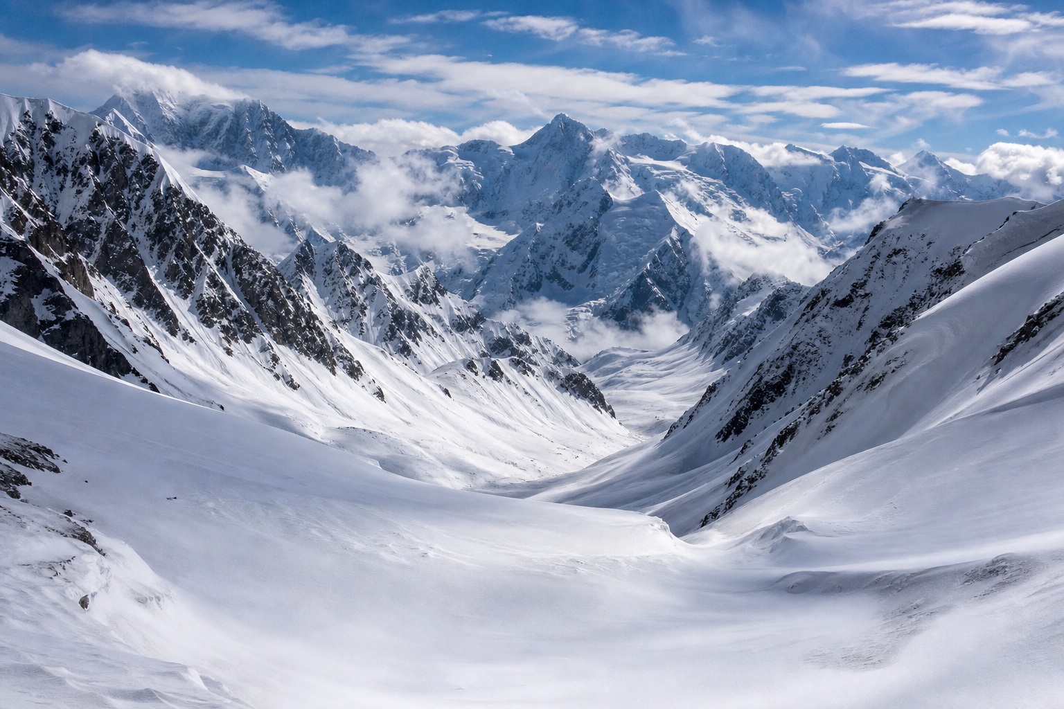 Snow-covered mountain pass between rugged peaks under a cloudy blue sky.