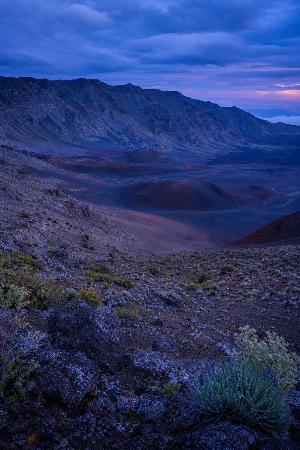 Blue hour view of a volcanic crater basin with rugged basalt slopes and rocky foreground.