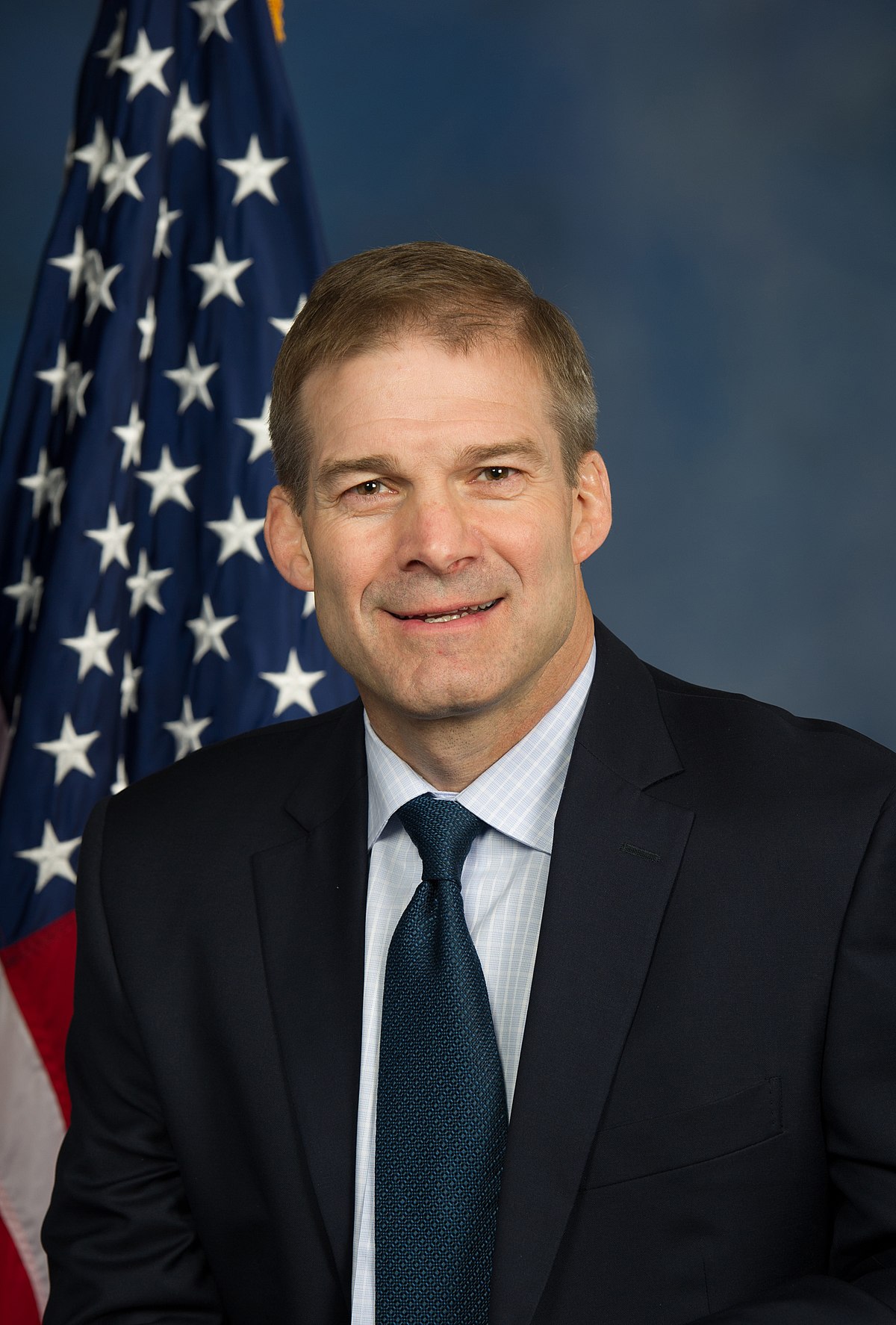 A smiling man in a dark suit and tie posed beside an American flag.