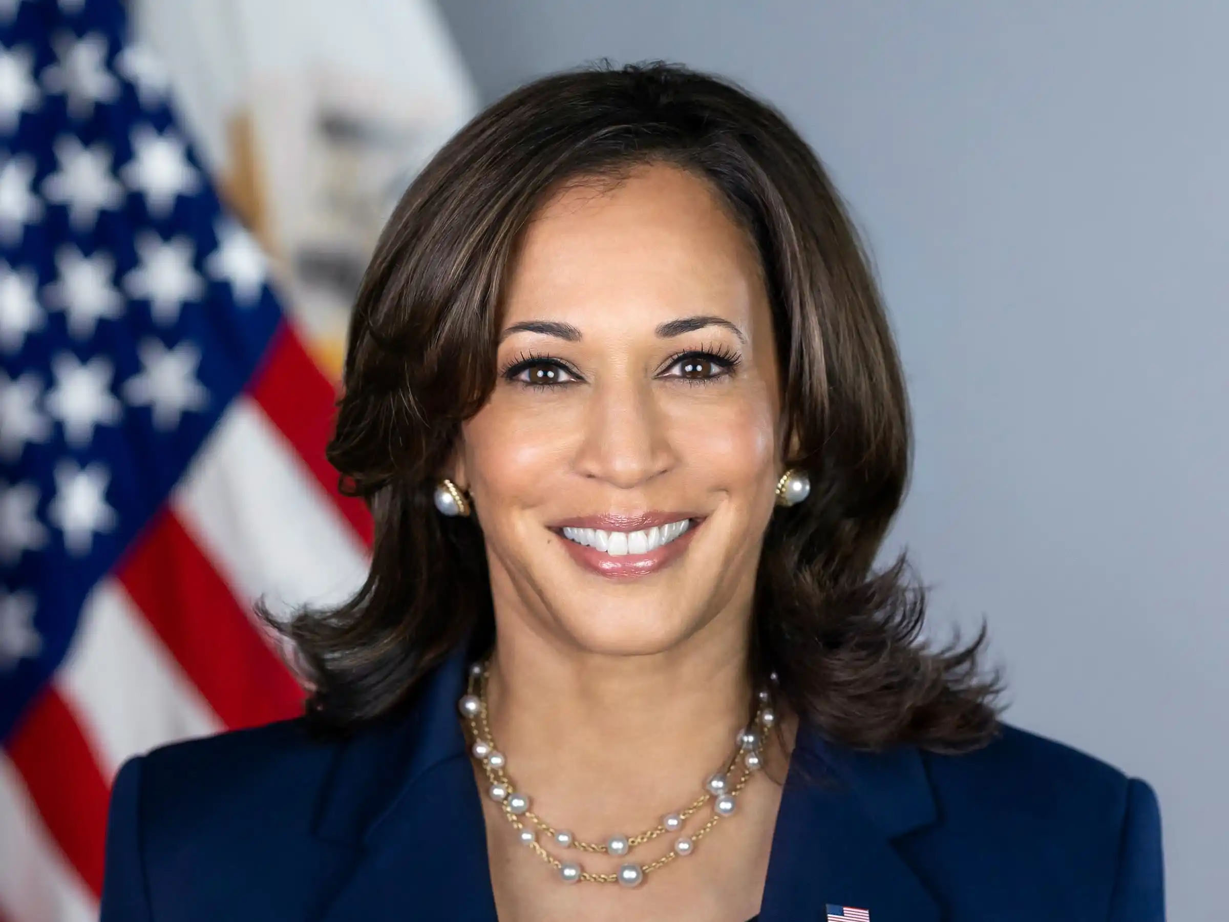 Smiling official portrait of Kamala Harris in a dark blazer with an American flag background
