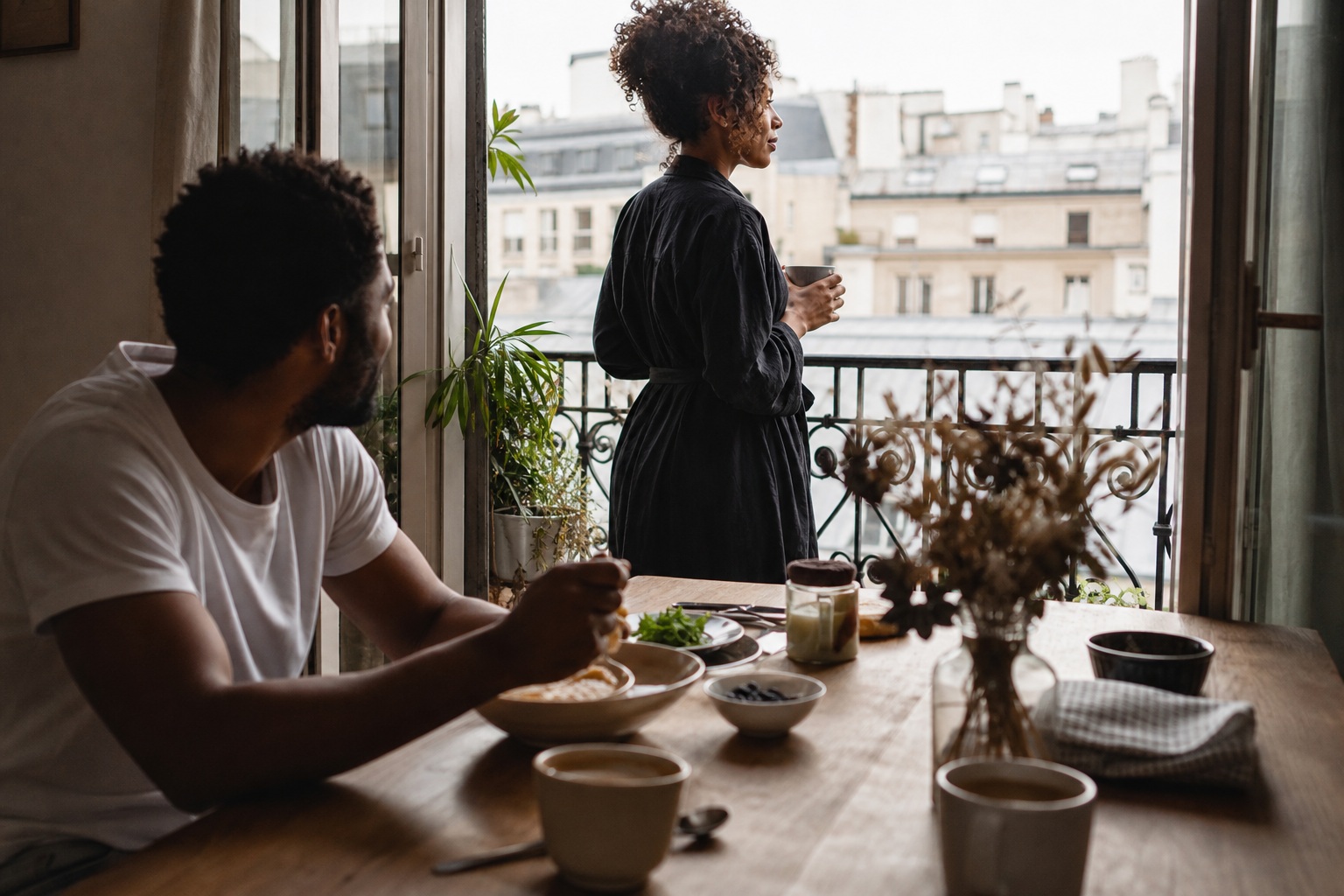 Two people in an apartment kitchen with breakfast on a table by a large window.