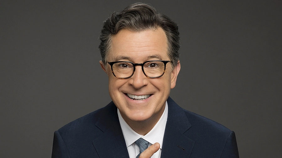 Smiling Stephen Colbert in glasses and a dark suit on a gray background.