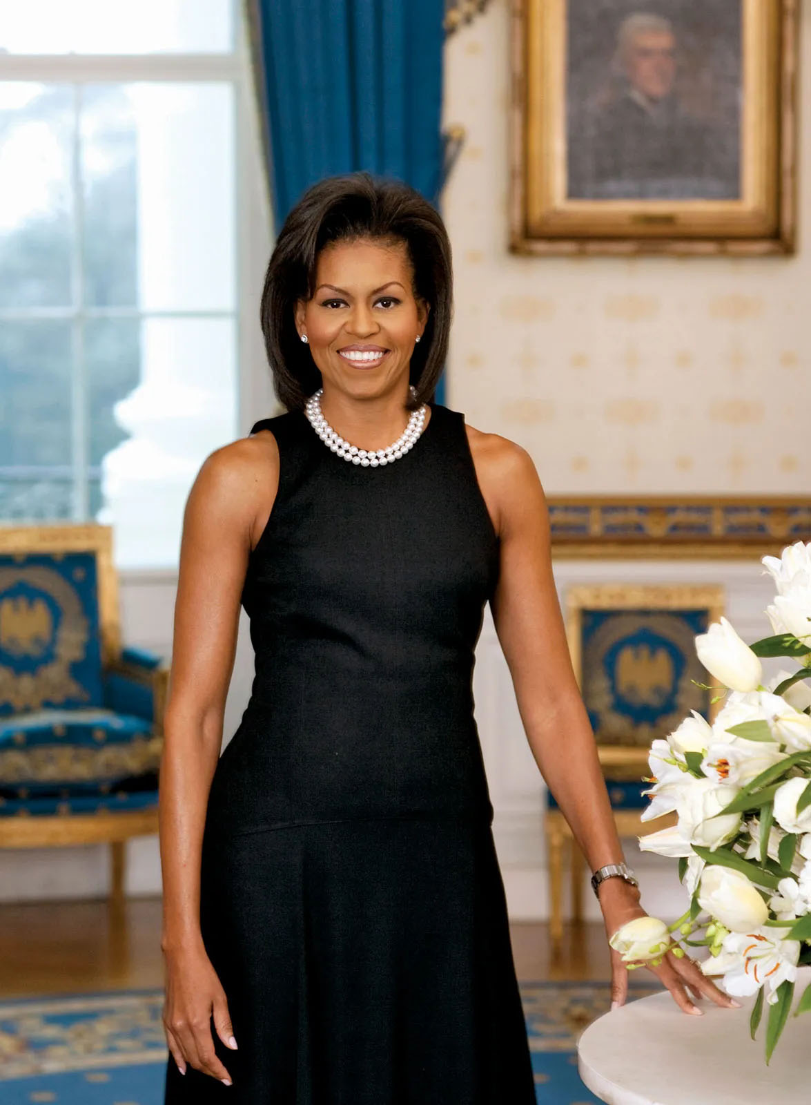 Michelle Obama standing in a black sleeveless dress beside a table of white flowers.