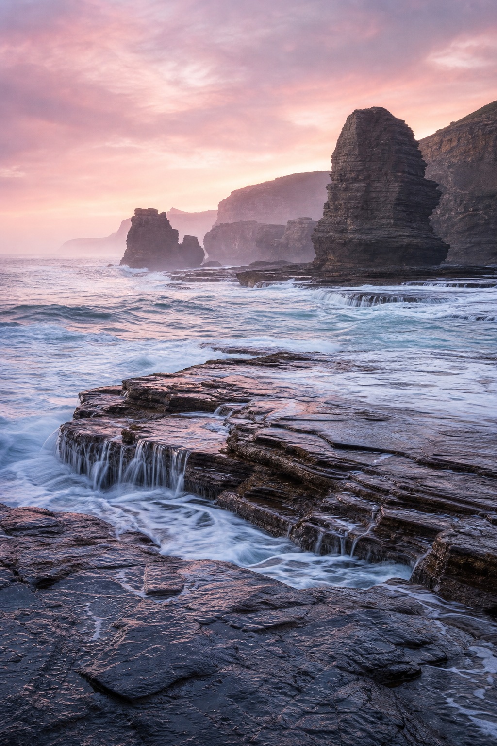 Sea stacks rising above a rocky coast with waves washing over a flat rock shelf at pink dawn.